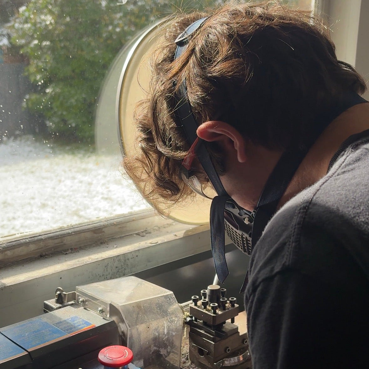 Adult man (Blake) working on a ring using a metal lathe with a window showing a tree in the background