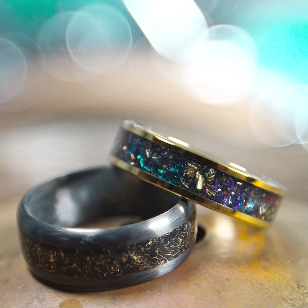 Two rings, one carbon fiber with cymbal shavings and a gold plated tungsten ring with multi color shifting pigments and cymbal shavings, the rings rest on a drum cymbal behind a blurred background