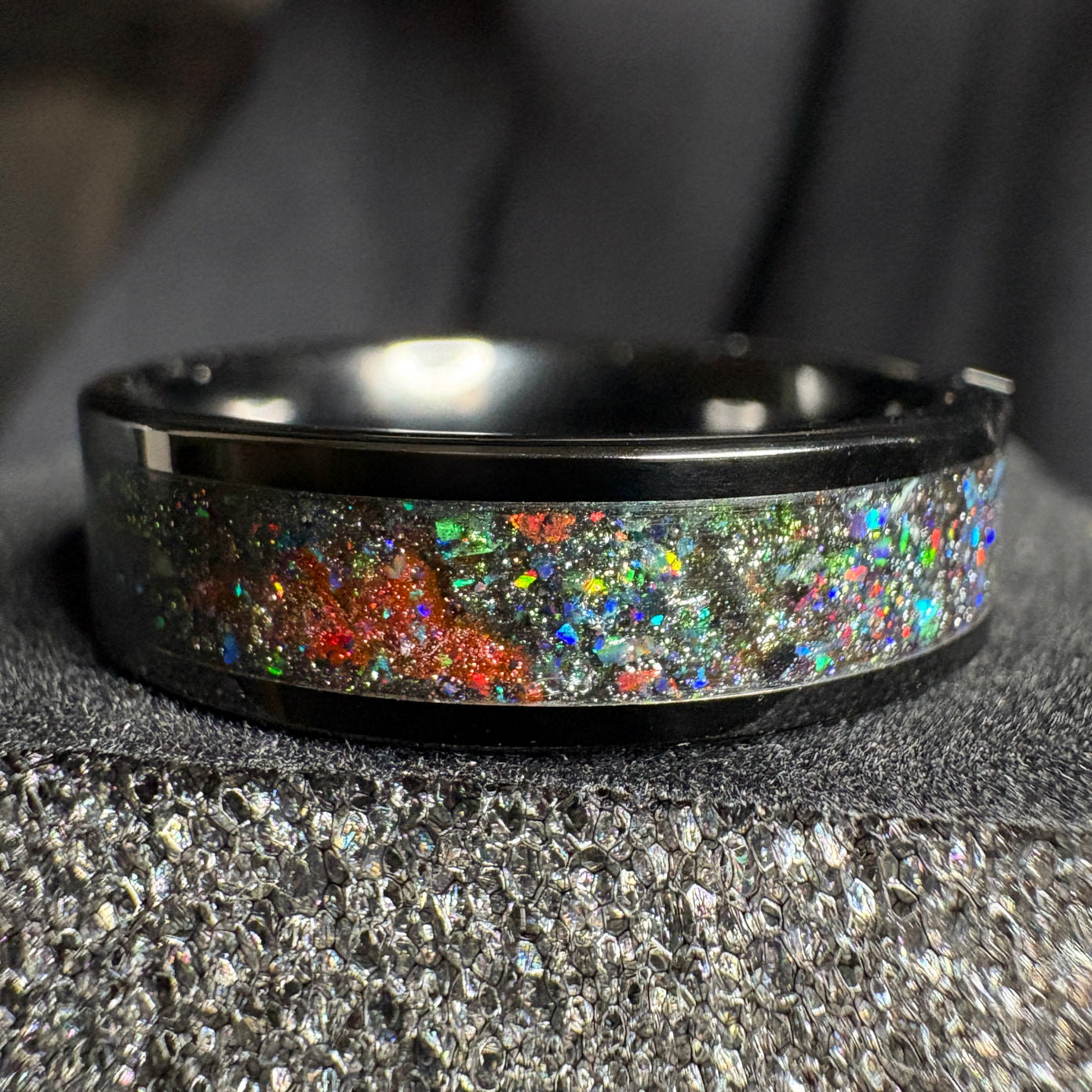 Black plated tungsten ring with multi color space themed shifting pigment powders and synthetic opals on a grey surface