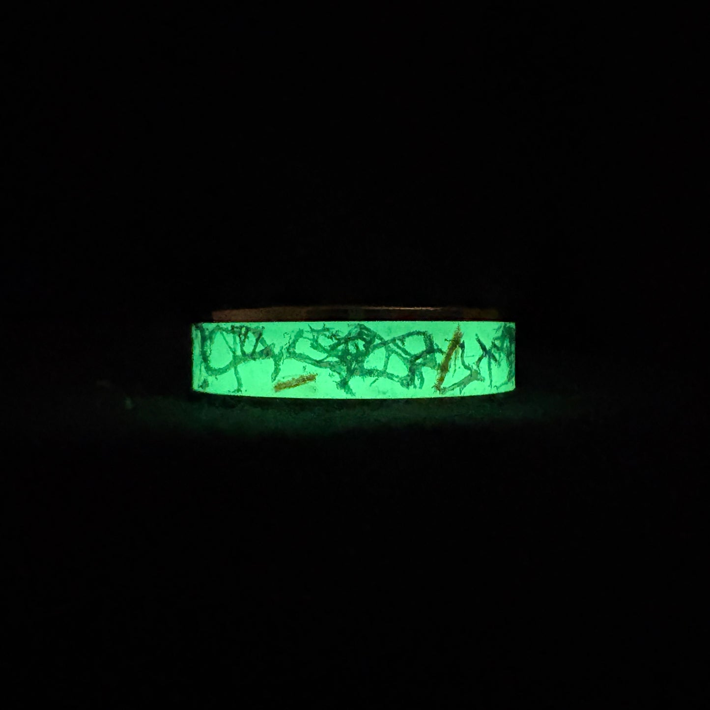 Gold plated tungsten ring with green glow in the dark powder, dried moss, and twigs inside in the dark glowing green