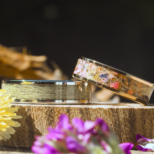 Two nature themed rings, one with dried flowers, one with moss, on a wooden block surrounded by dried flowers
