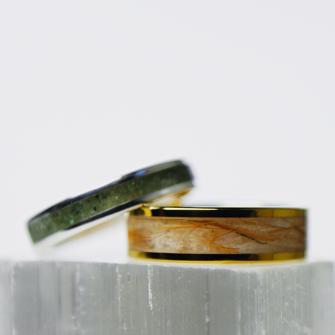 Two rings, one with cat fur, one with sand and gemstones, sitting on a white surface