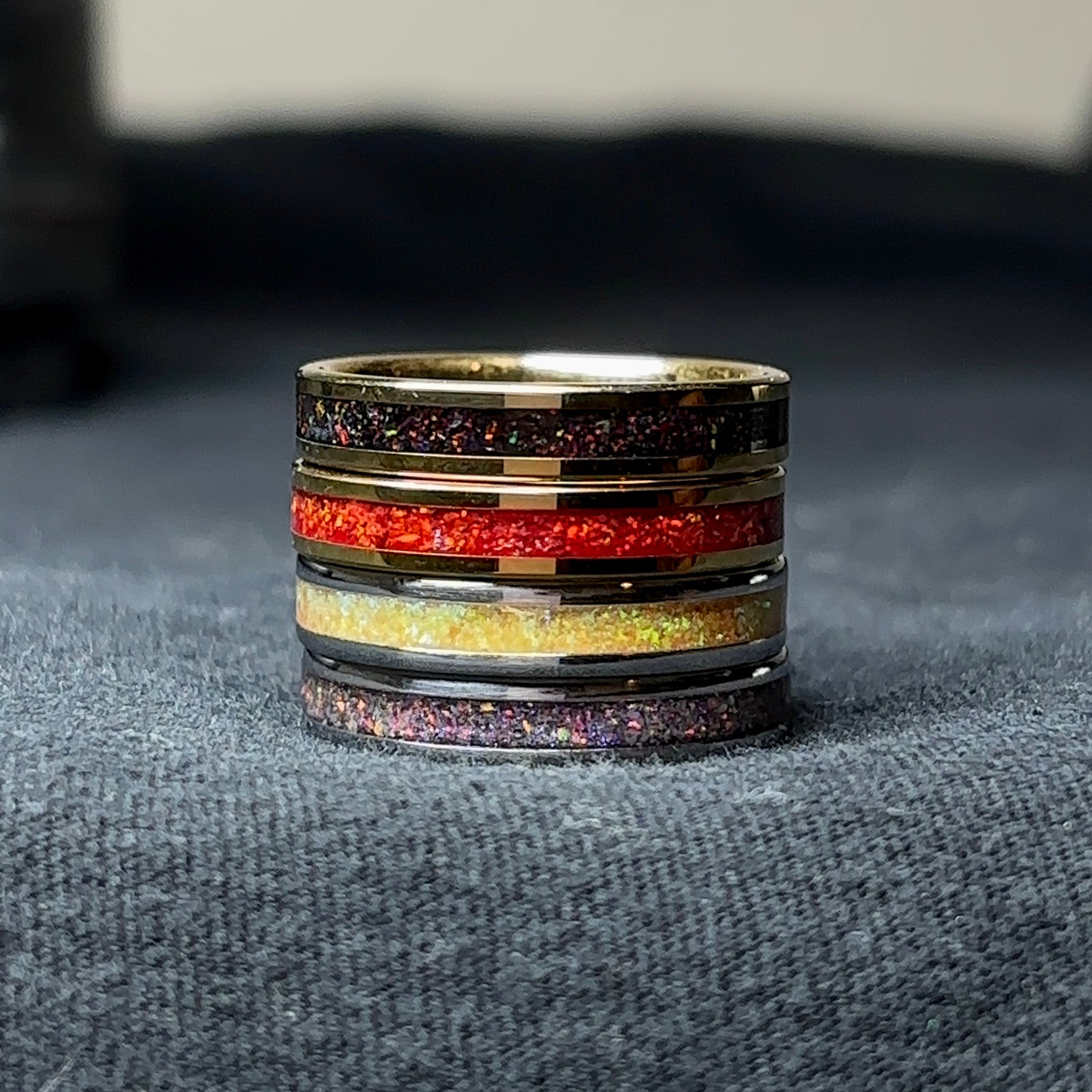 4 gold plated tungsten rings, one with black opal, one with red opal, one with yellow opal, one with purple opal, stacked on top of one another on a black surface